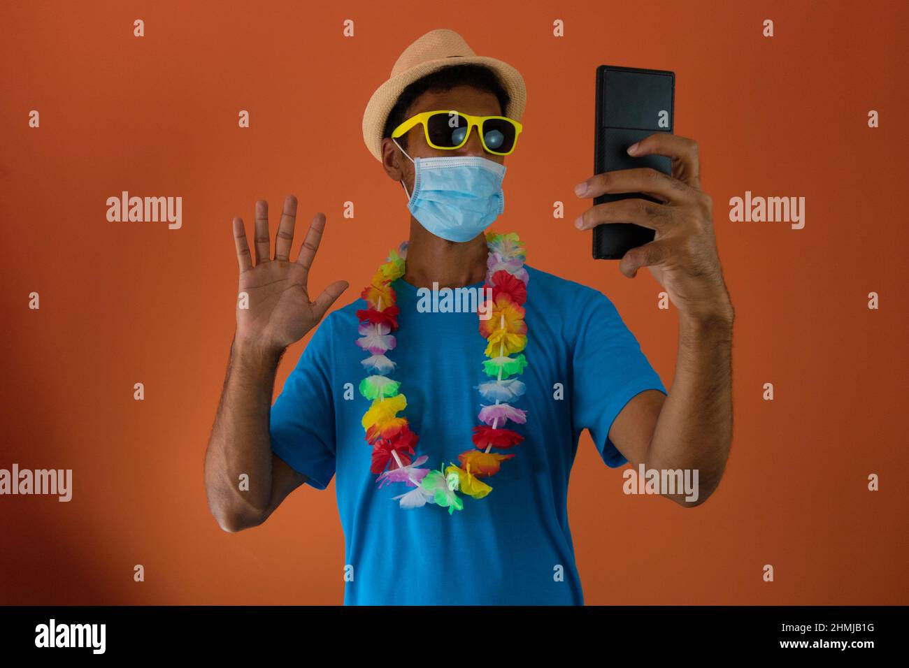 Homme noir en costume de carnaval et masque pandémique tenant un téléphone portable isolé sur fond orange. Banque D'Images