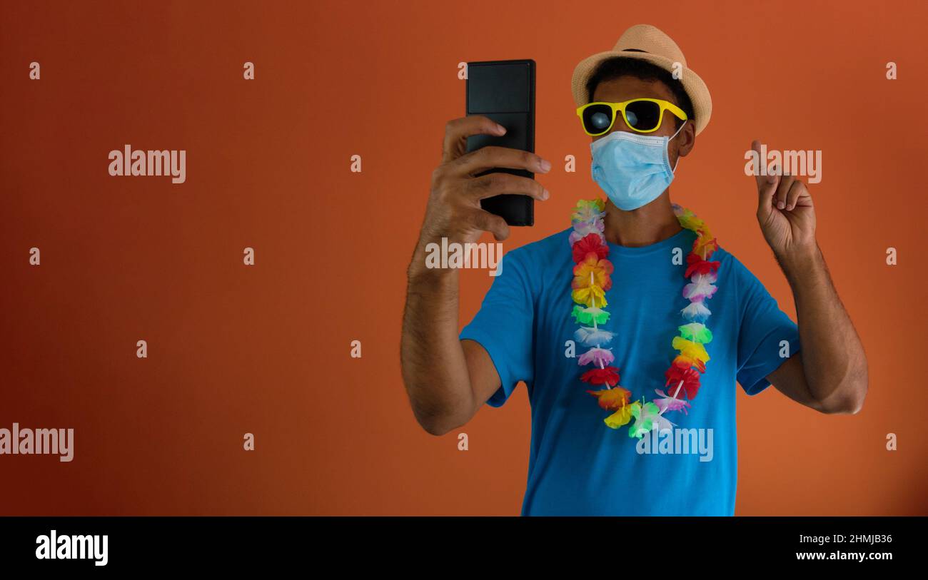 Homme noir en costume de carnaval et masque pandémique tenant un téléphone portable isolé sur fond orange. Banque D'Images