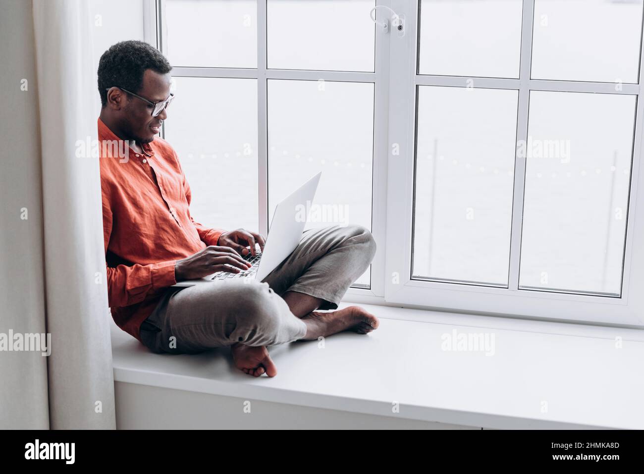 Un homme afro-américain séduisant en vêtements et lunettes décontractés surfe sur Internet sur un ordinateur portable moderne assis sur le seuil de la fenêtre à la maison Banque D'Images