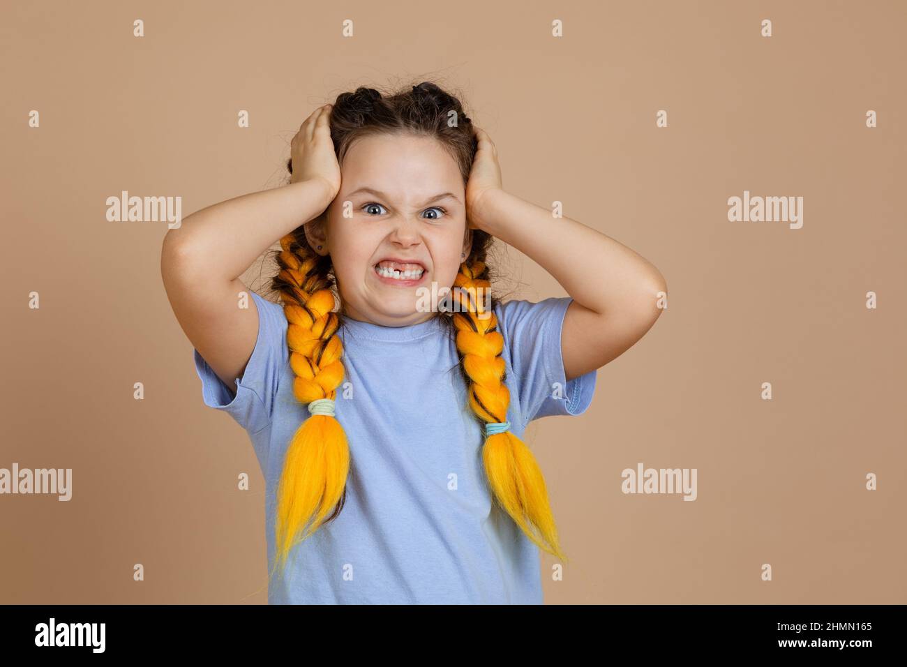 Colère offensé contrarié caucasien petite fille tenant la tête grinning dents regardant la caméra ayant kanekalon braides de couleur jaune dans le t-shirt bleu sur Banque D'Images