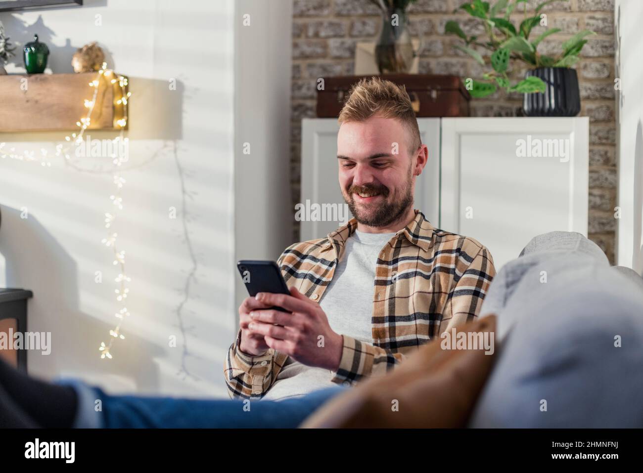 Vue de face d'un jeune homme allongé sur son canapé dans son salon relaxant, il utilise son smartphone. Banque D'Images
