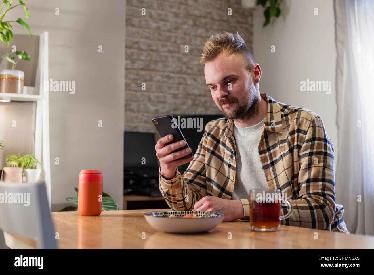 Vue latérale d'un jeune homme assis à une table dans son salon relaxant, il utilise son smartphone et il a une assiette de pâtes à proximité. Banque D'Images