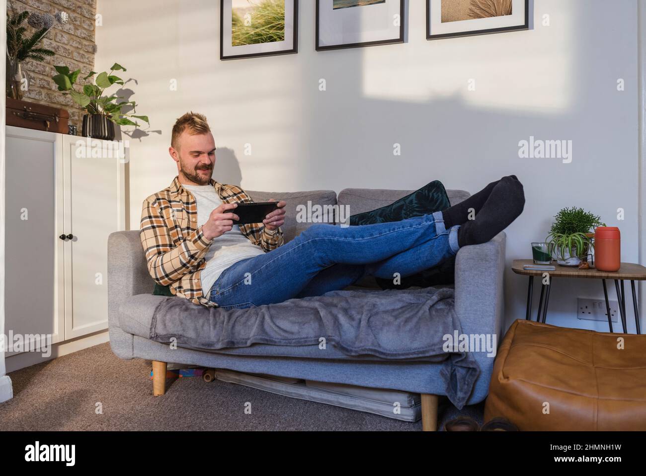 Vue latérale d'un jeune homme allongé sur son canapé dans son salon reposant, il utilise son smartphone. Banque D'Images