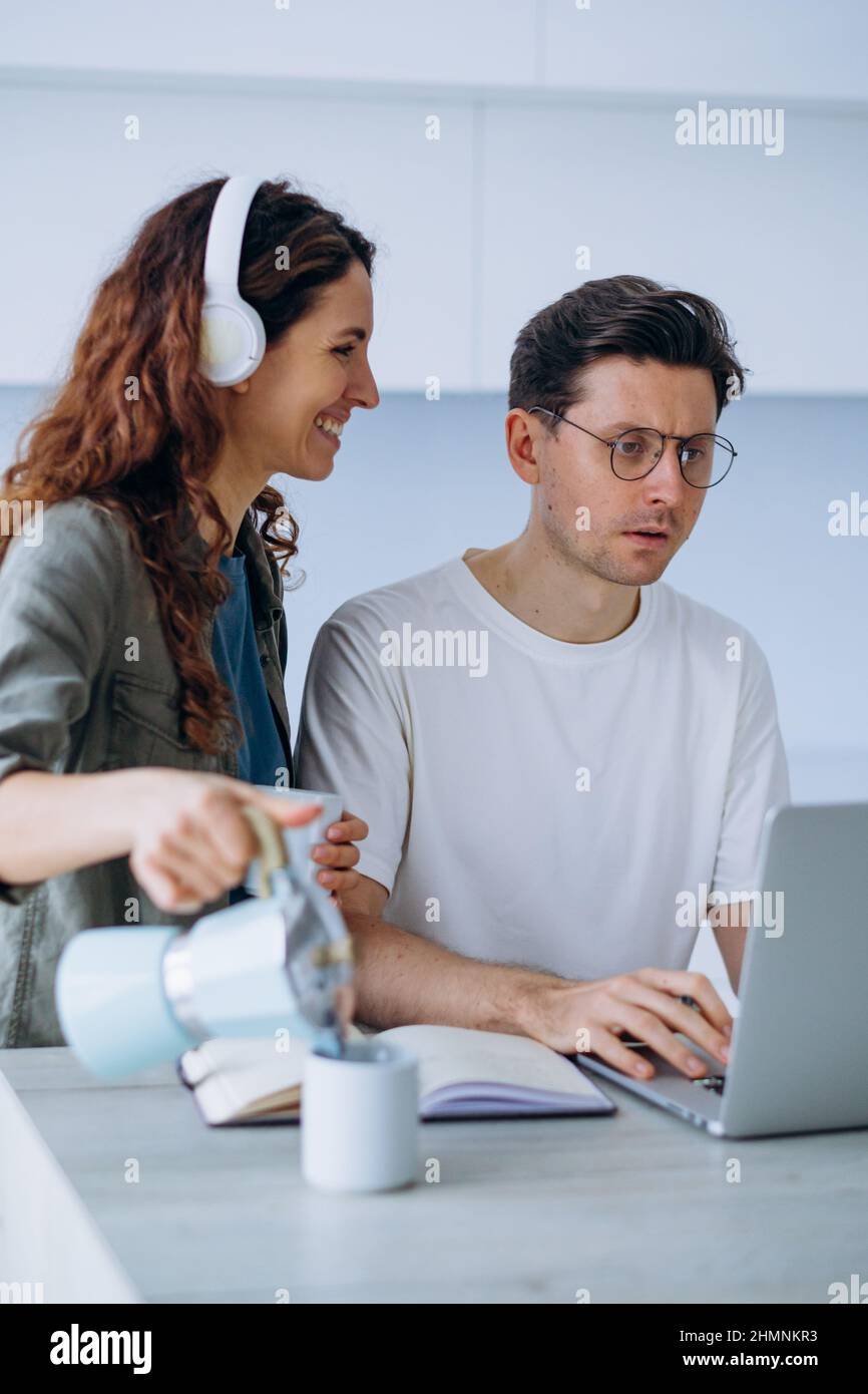 Une femme aux cheveux longs et bouclés sourit dans un casque et verse du café dans une tasse pour un mari stressé travaillant sur un ordinateur portable et résolvant les problèmes de travail Banque D'Images