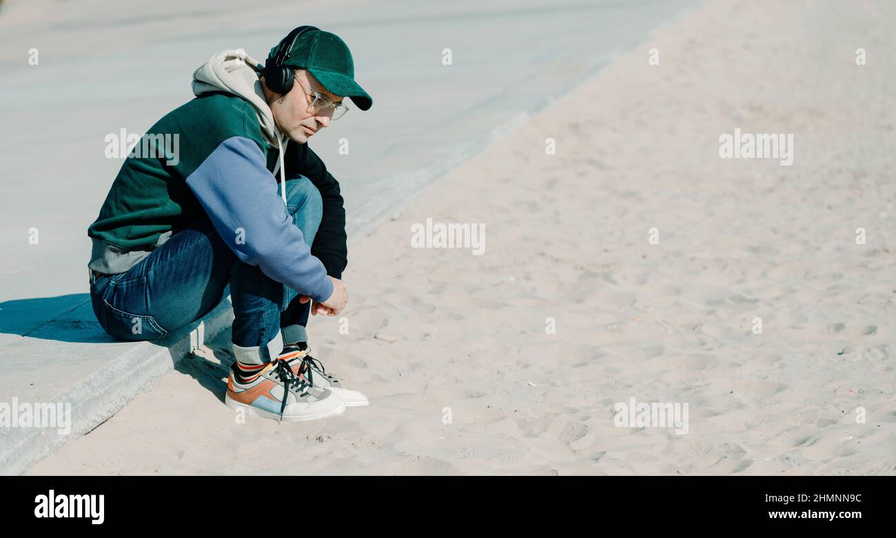 un beau homme d'âge moyen, portant des vêtements modernes et décontractés, écoute de la musique à l'aide d'un casque assis sur la promenade, à côté du sable d'un Banque D'Images