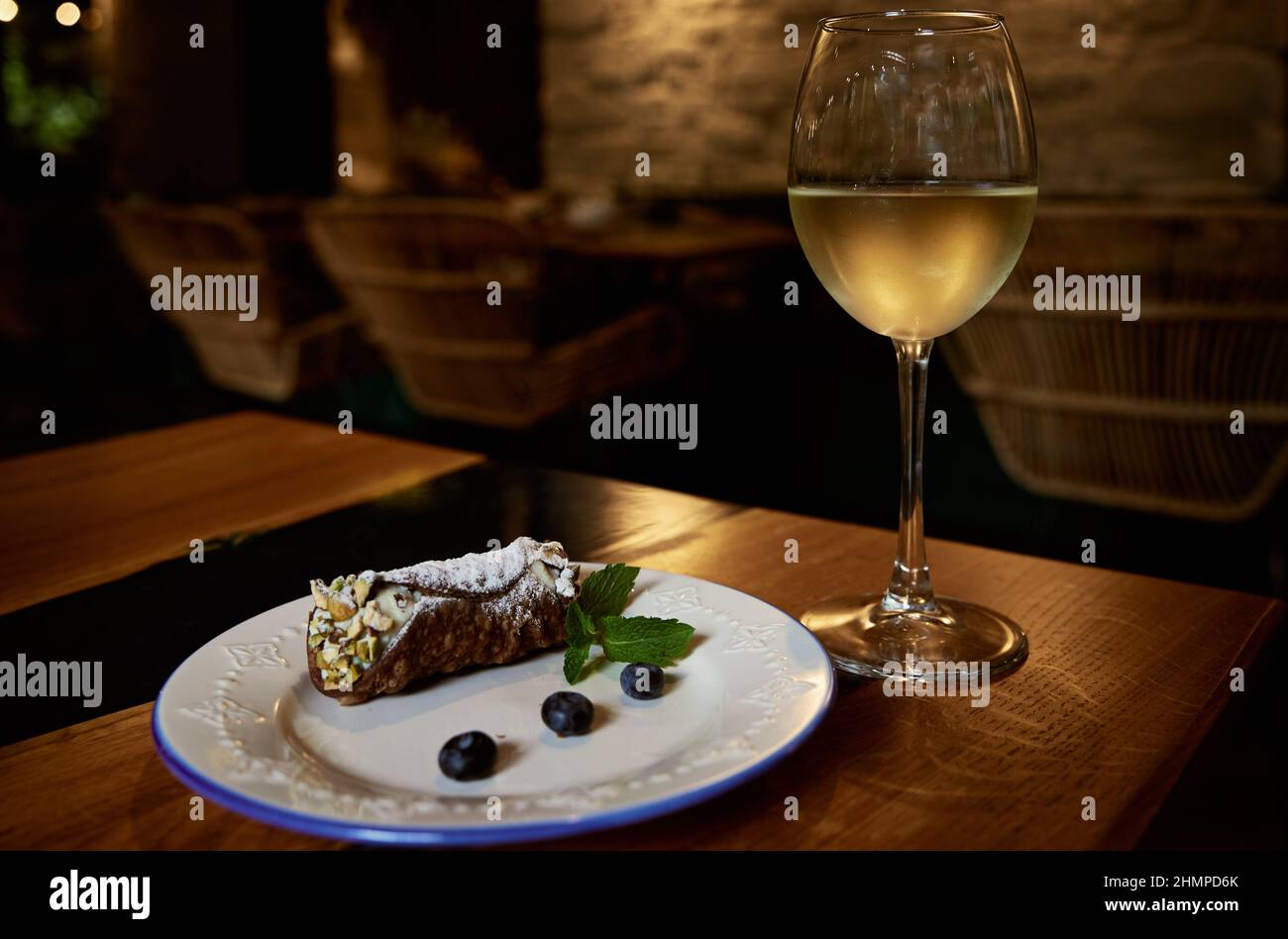 Des cannoli siciliens roulent avec des bleuets sur une plaque de céramique blanche et un verre de vin blanc misté sur une table de restaurant avec un fond flou. Banque D'Images