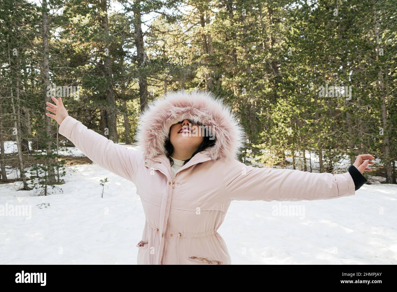 Femme souriante portant un manteau d'hiver avec capuche en fausse fourrure debout dans une forêt enneigée avec ses bras étirés, Espagne Banque D'Images