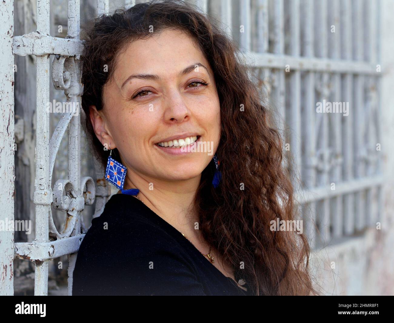 Belle positive jeune mexicaine caucasienne femme avec de longues boucles sourires devant un grill blanc de fenêtre en fer forgé et regarde le spectateur. Banque D'Images
