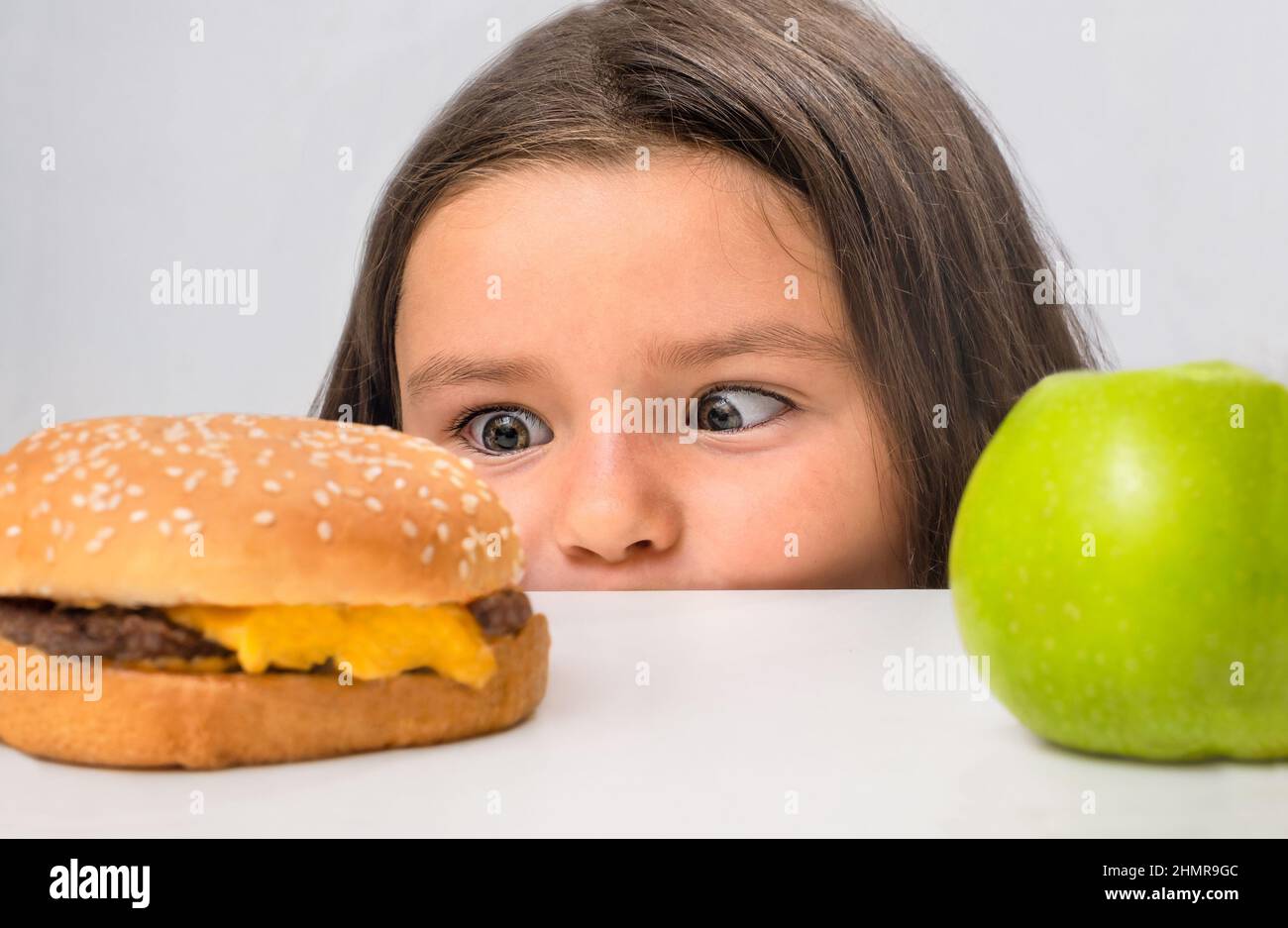 L'enfant regarde derrière la table et choisit entre un hamburger et une pomme Banque D'Images