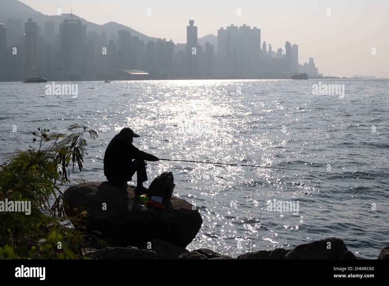 Silhouette d'homme assis sur le rocher, pêche au coucher du soleil au port de Victoria avec paysage urbain de Hong Kong derrière Banque D'Images