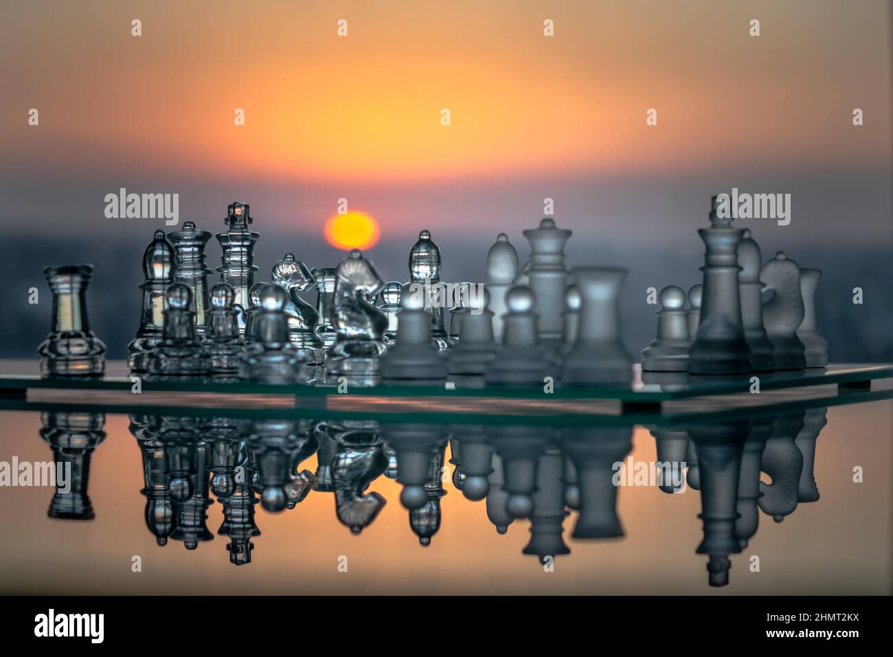 Un jeu d'échecs en verre placé sur une table en verre vue du côté avec un reflet horizontal et un fond de couleur orange avec un cercle solaire jaune coucher de soleil. Banque D'Images