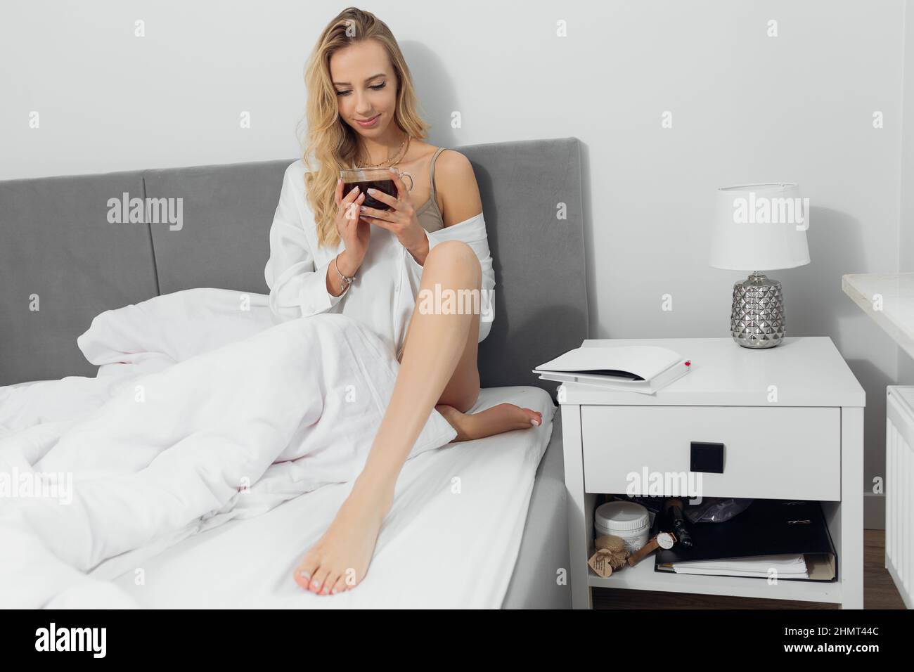 Portrait d'une jeune femme blonde pieds nus. Porter des vêtements blancs. Auto-soin et auto-étude.boire du café dans le lit. Banque D'Images