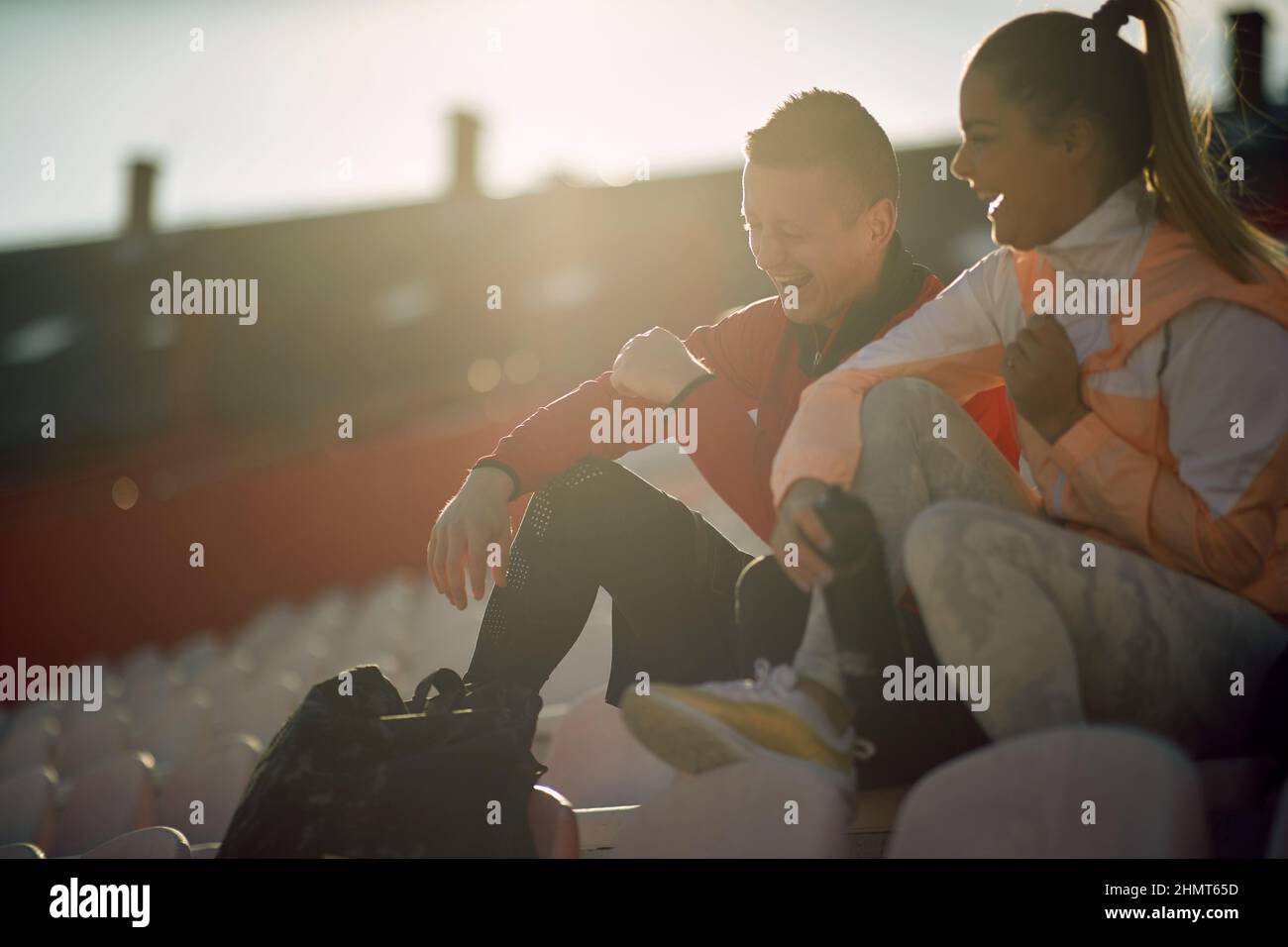 un jeune couple caucasien adulte riant à voix haute, assis à la tribune du stade dans des vêtements de sport. Banque D'Images
