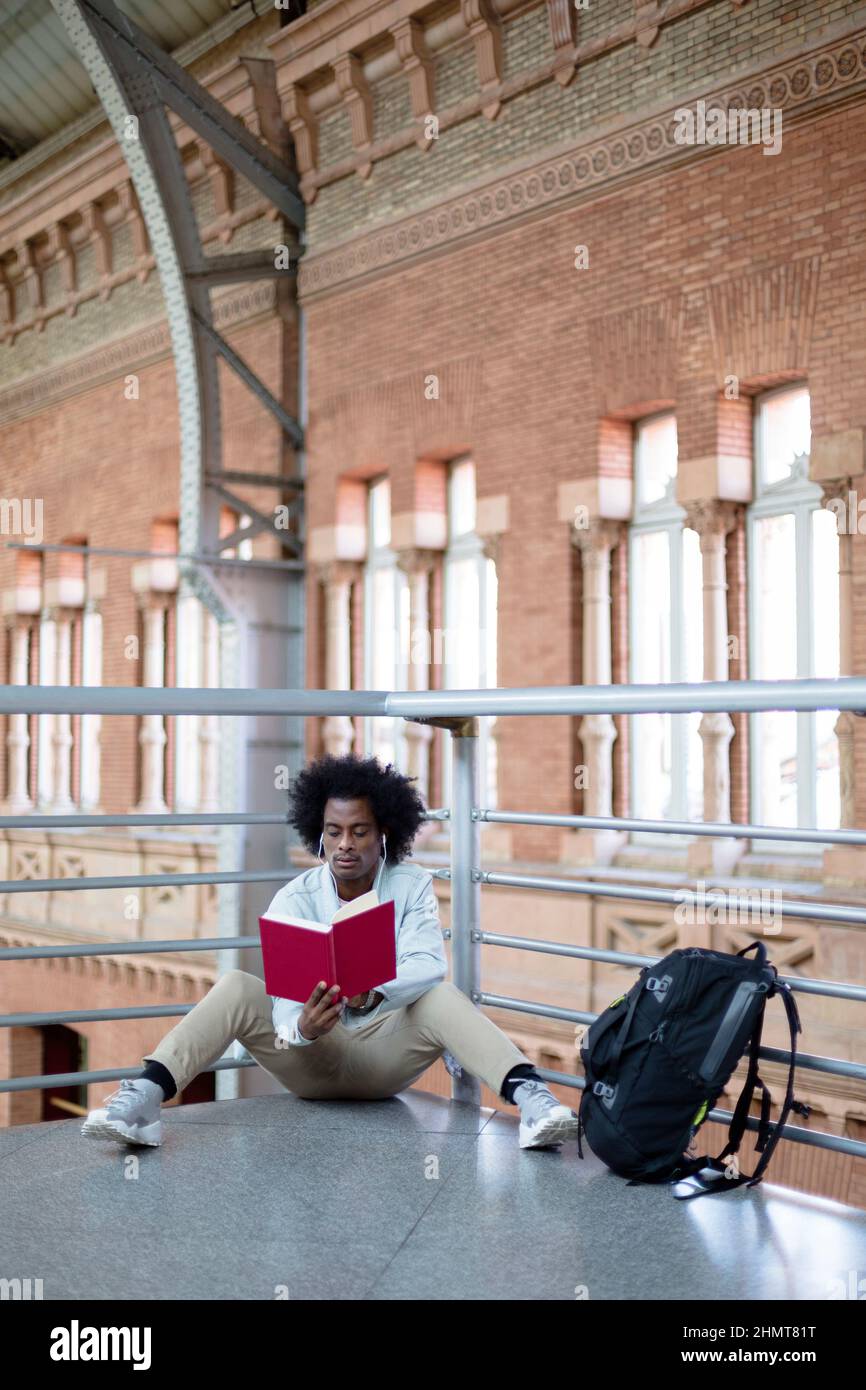 Homme afro-américain avec une coiffure afro et des vêtements décontractés lisant un livre à la gare. Concept de voyage et de style de vie. Espace pour le texte. Banque D'Images