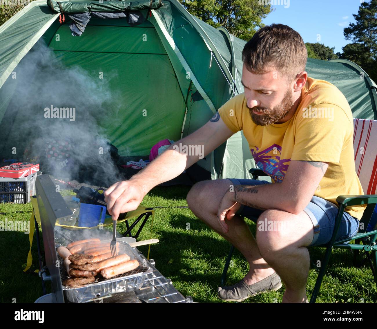 Homme campant et utilisant un barbecue jetable pour cuire des hamburgers et des saucisses Banque D'Images
