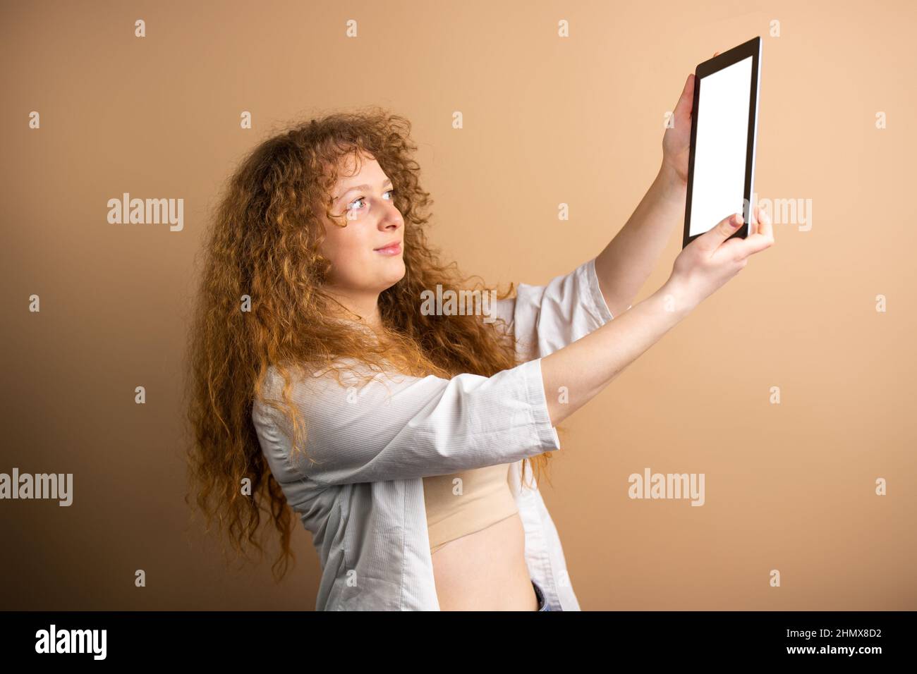 Photo de vue latérale d'une jeune femme à tête rouge tenant dans ses mains tablette et allant faire selfie sur fond d'or. Banque D'Images
