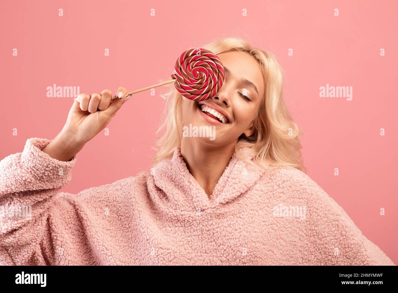 Jeune femme joyeuse tenant des bonbons sur le bâton, couvrant un oeil avec le lollipop et souriant, s'amusant sur fond rose Banque D'Images