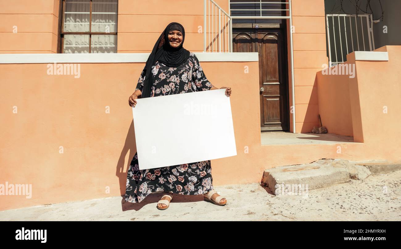 Joyeuse femme musulmane tenant un écriteau vierge et souriant à la caméra tout en se tenant devant sa maison pendant la journée. woma d'âge moyen et confiant Banque D'Images