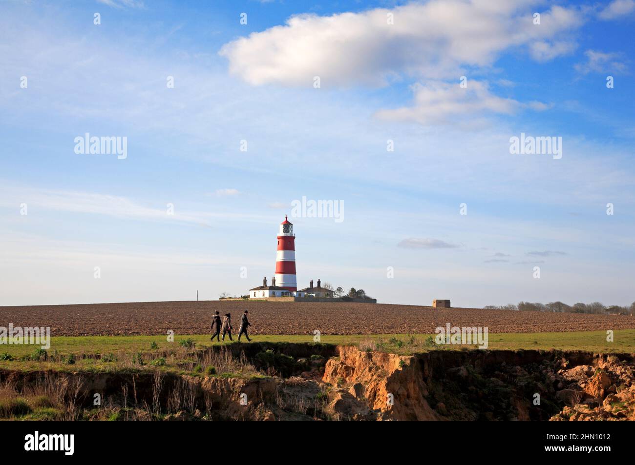 Trois personnes marchant sur un sentier au sommet d'une falaise en érodant les falaises avec un phare en arrière-plan à Happisburgh, Norfolk, Angleterre, Royaume-Uni. Banque D'Images