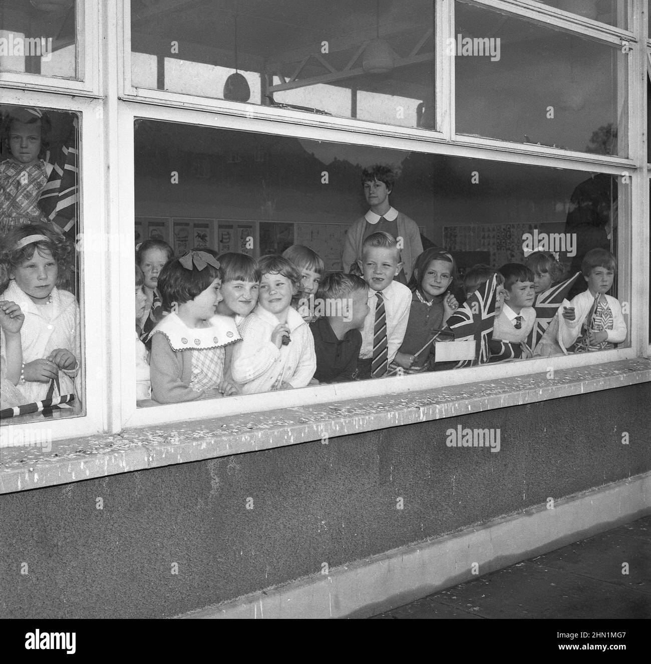 1965, des garçons et des filles de l'école primaire, historiques et excités, regardant par la fenêtre de leur salle de classe, certains avec des drapeaux Union Jack en main, Angleterre, Royaume-Uni. Banque D'Images