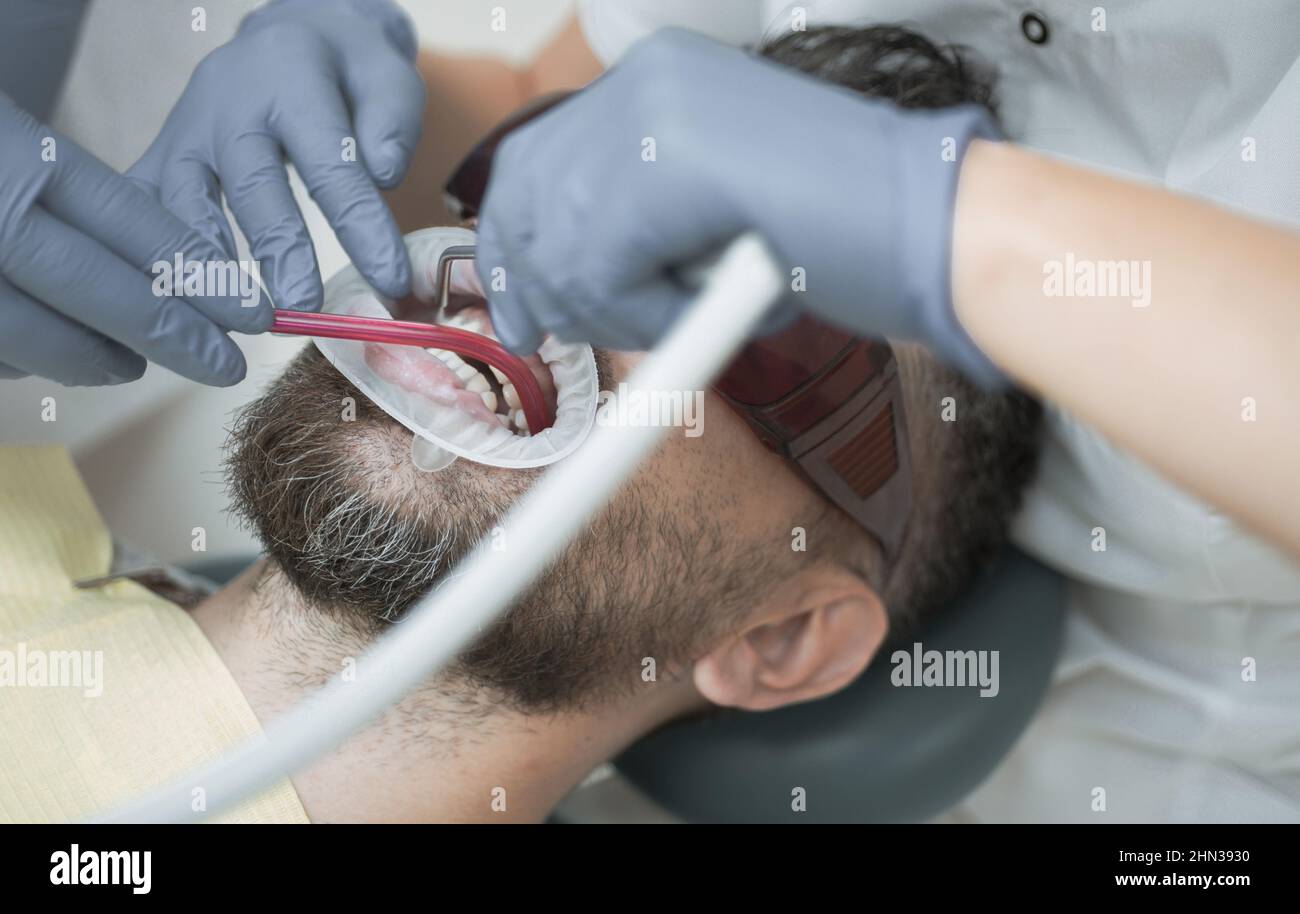 Jeune homme choisissant la couleur des dents chez le dentiste, gros plan. Dentiste femme vérifiant les dents du patient avec miroir dans la clinique dentaire moderne. Bel homme à Banque D'Images