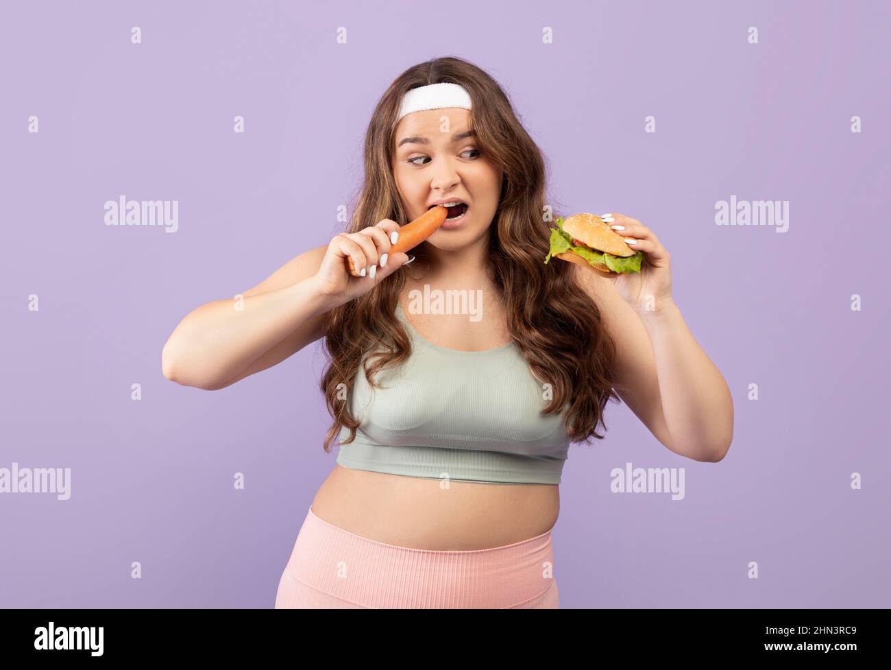 Petite fille européenne pensive et affamée de vêtements de sport mange la carotte et regarde le hamburger Banque D'Images