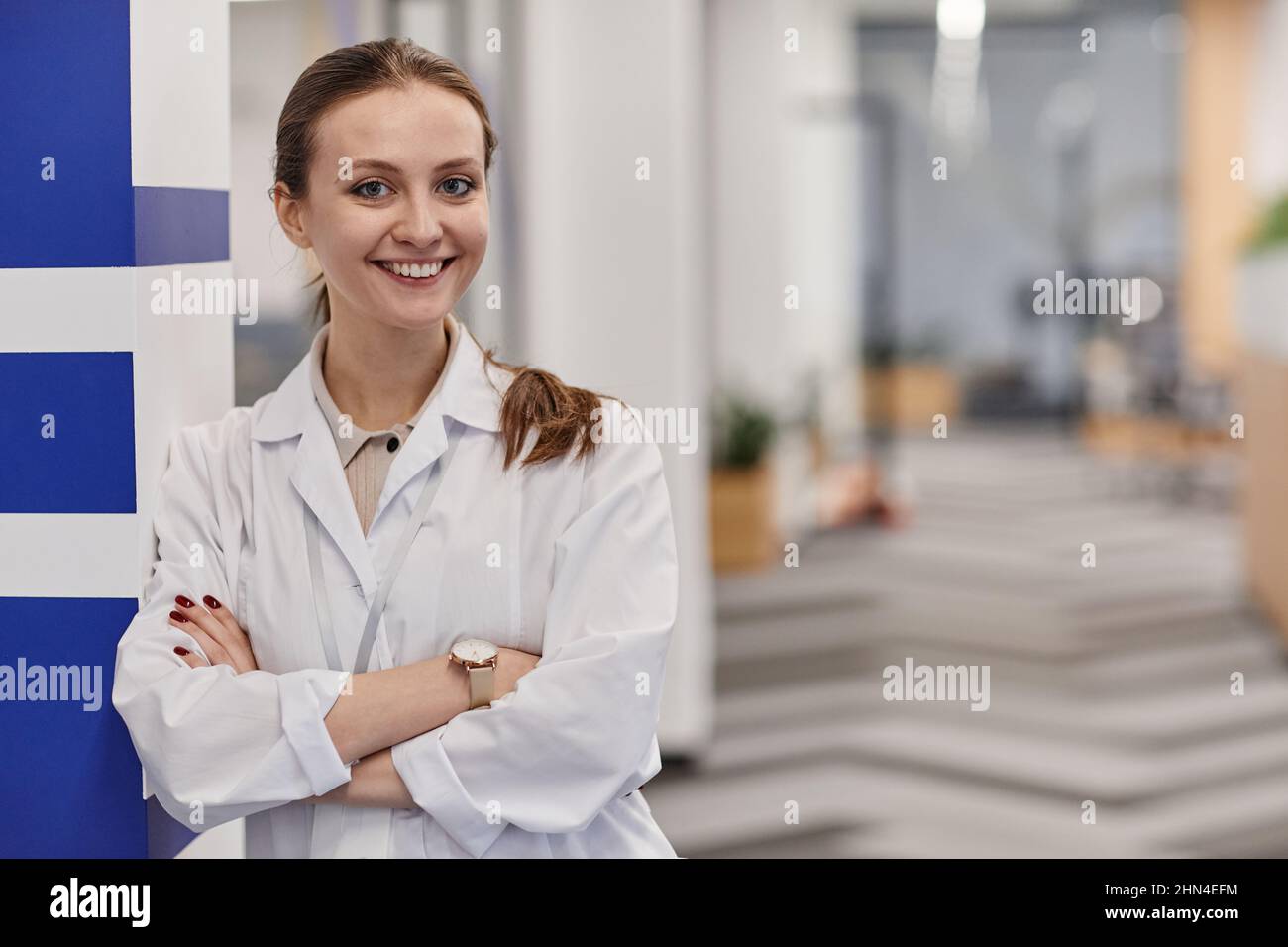 Jeune médecin-hygiéniste heureuse portant un manteau de laboratoire croisant les bras par la poitrine tout en se tenant dans le couloir des cliniques contemporaines Banque D'Images