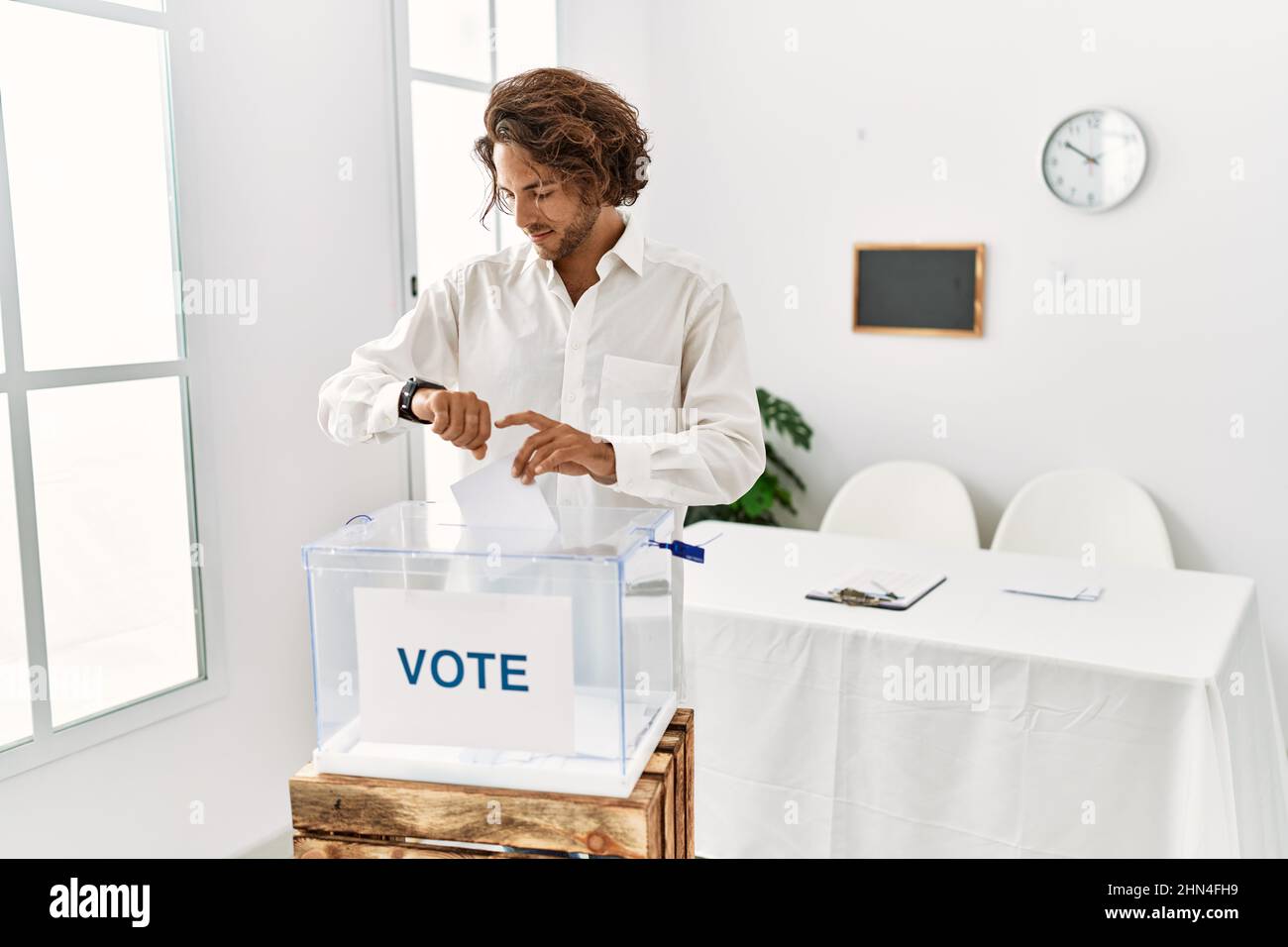 Jeune hispanique qui vote une enveloppe dans les urnes vérifiant l'heure sur la montre de poignet, détendu et confiant Banque D'Images