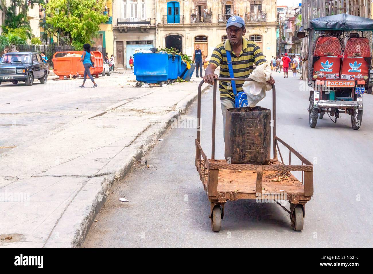 Un homme afro-caribéen pousse un chariot dans une rue de ville. Un bicitaxi et une poubelle sont vus en arrière-plan. Banque D'Images