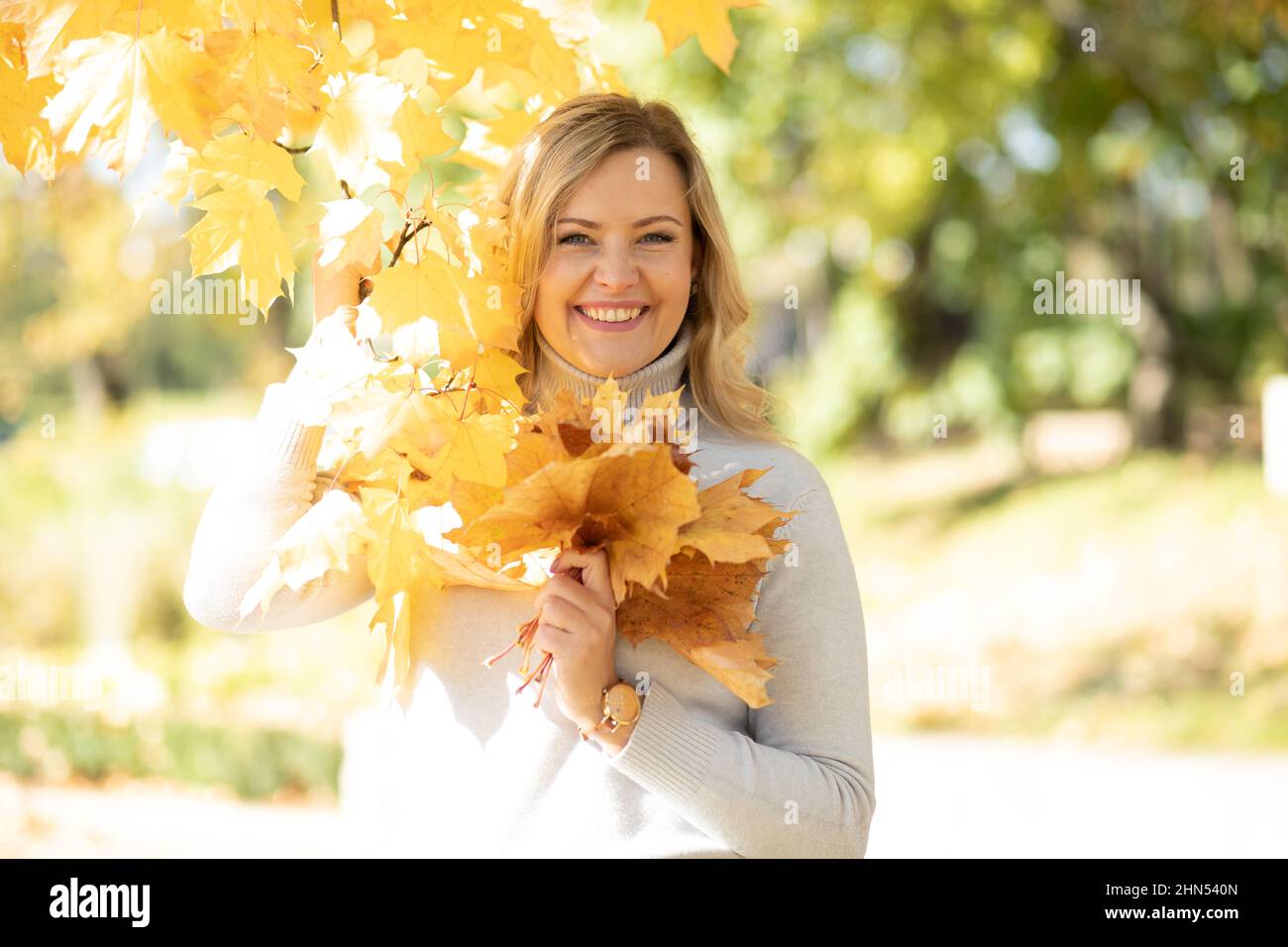 Belle femme d'âge moyen souriante avec de longs cheveux ondulés justes dans le chandail à col roulé tenir un bouquet de feuilles jaunes tombées. Banque D'Images