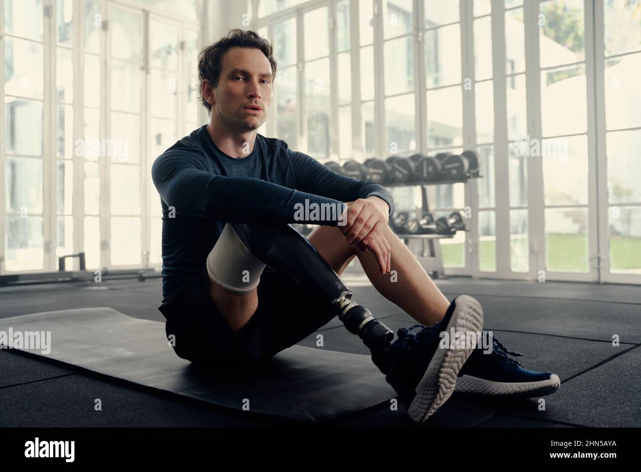 Portrait d'un athlète paralympique masculin avec une jambe prothétique assise sur un tapis regardant dans la caméra avec une expression sérieuse sur son visage. Banque D'Images