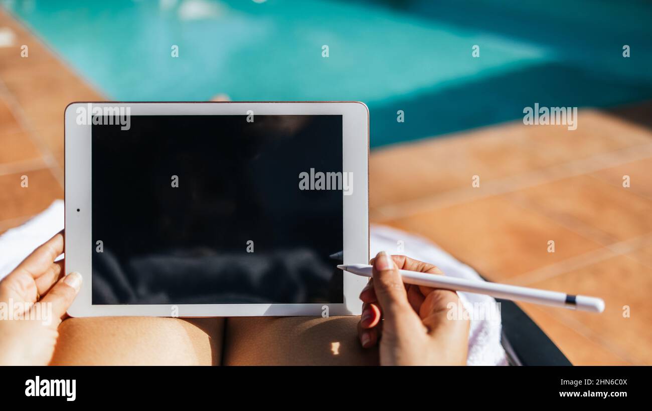 Gros plan d'une femme méconnue travaillant avec une tablette avec un stylo électronique à côté d'une piscine turquoise Banque D'Images