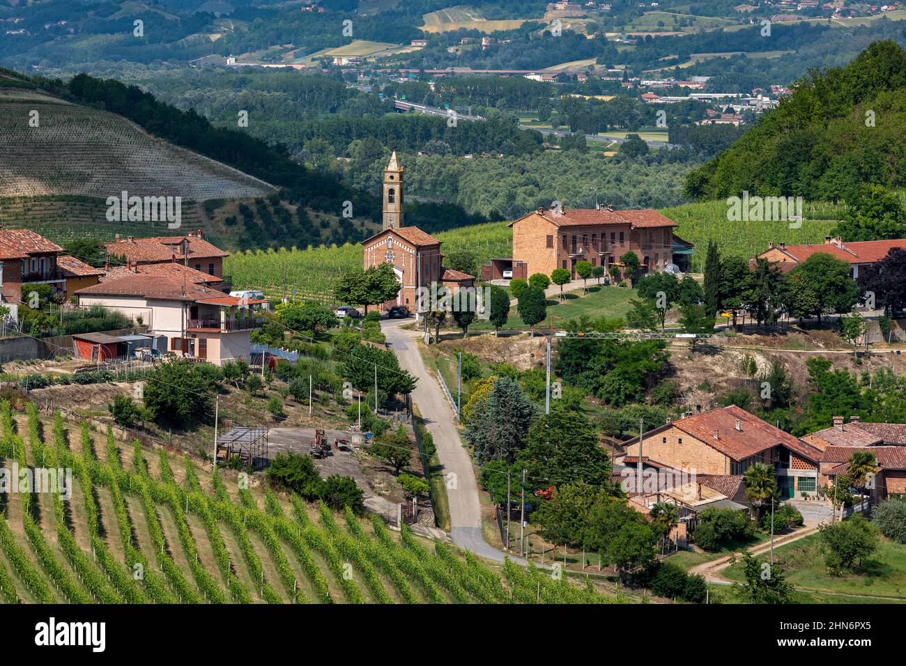 Vue sur la route rurale à travers le village et l'église paroissiale parmi les vignobles verts du Piémont, dans le nord de l'Italie. Banque D'Images