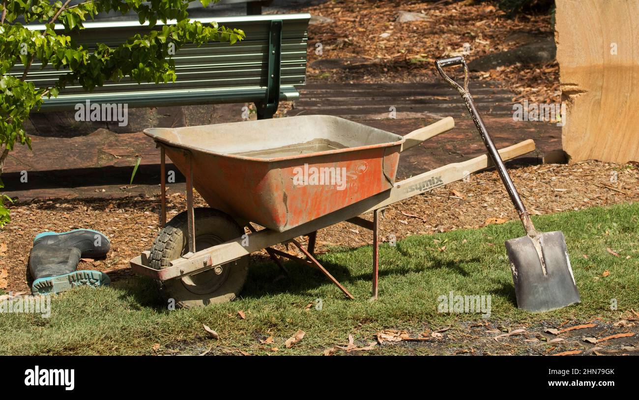 Brouette remplie de ciment avec une pelle penchée sur la poignée et des bottes en caoutchouc à proximité - équipement de travail sur le chantier de construction de jardin Banque D'Images