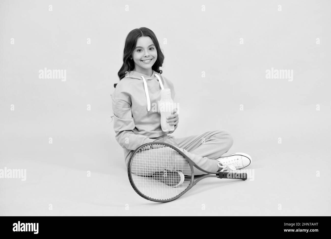 un enfant heureux s'assoit dans des vêtements de sport avec une bouteille d'eau de raquette de tennis sur fond jaune, équilibre aquatique Banque D'Images