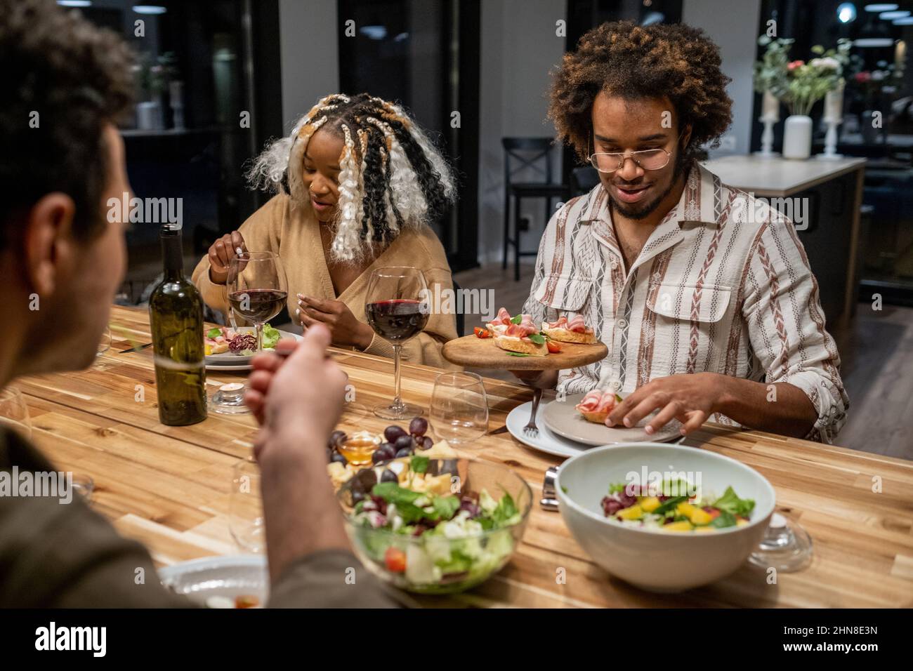Des jeunes heureux assis à la table à manger et manger de la salade et des hors-d'œuvre pendant le dîner à la maison Banque D'Images