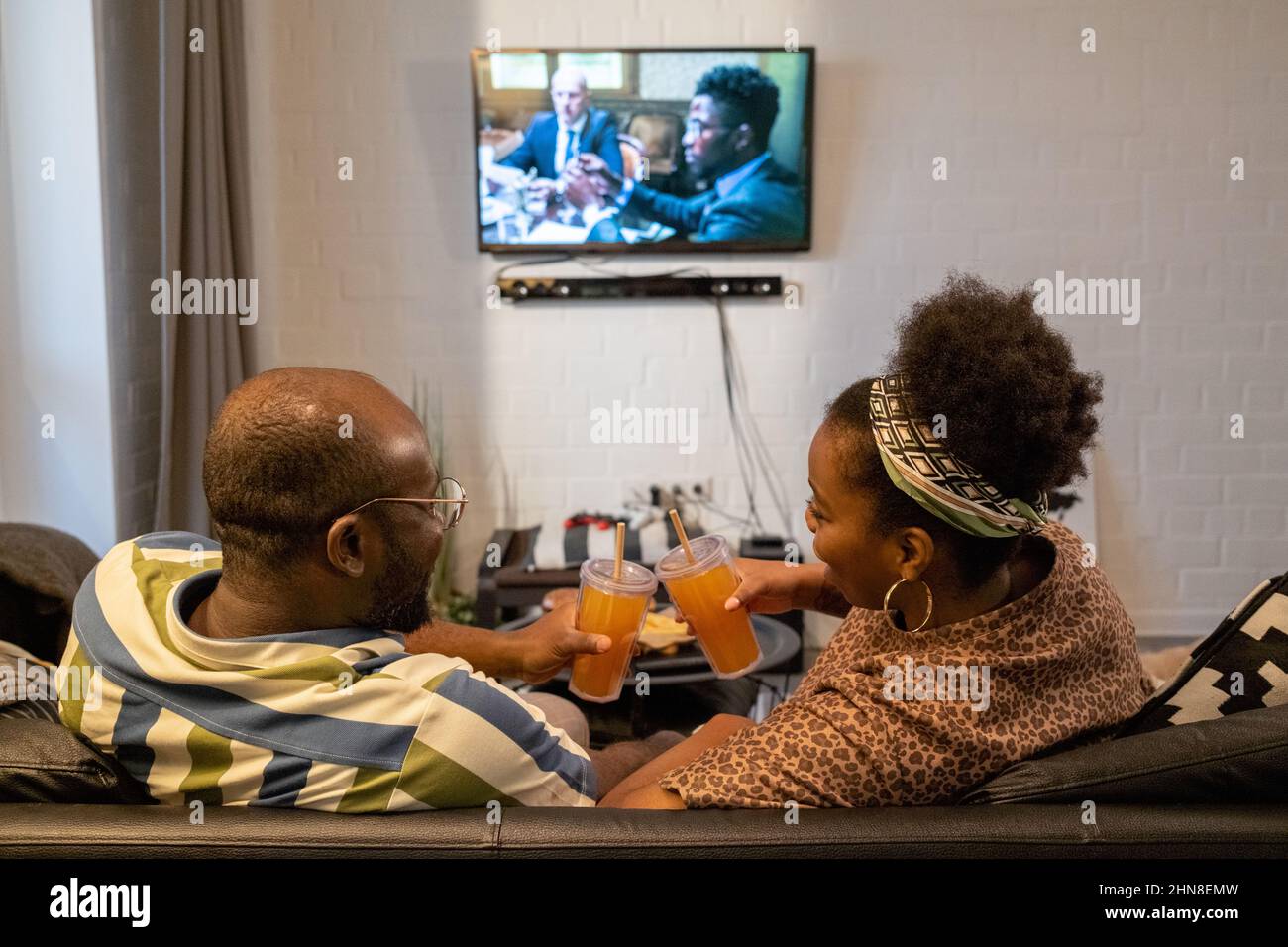 Vue arrière d'un couple africain en train de griller avec des verres de jus et de sourire tout en étant assis sur un canapé pendant que vous regardez le film à la maison Banque D'Images