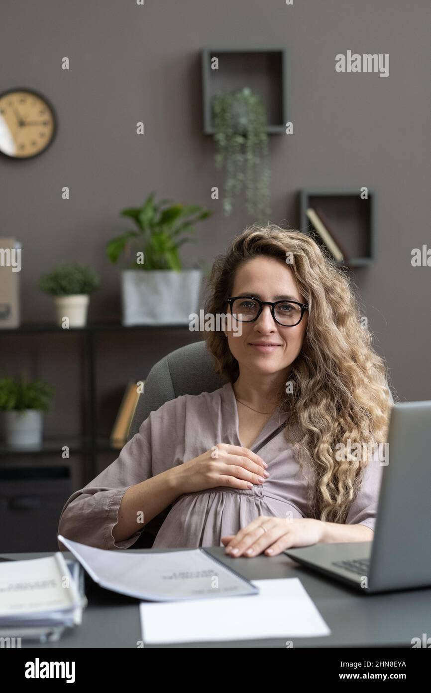 Portrait d'une jeune femme enceinte à lunettes regardant l'appareil photo tout en étant assise sur son lieu de travail avec un ordinateur portable au bureau Banque D'Images