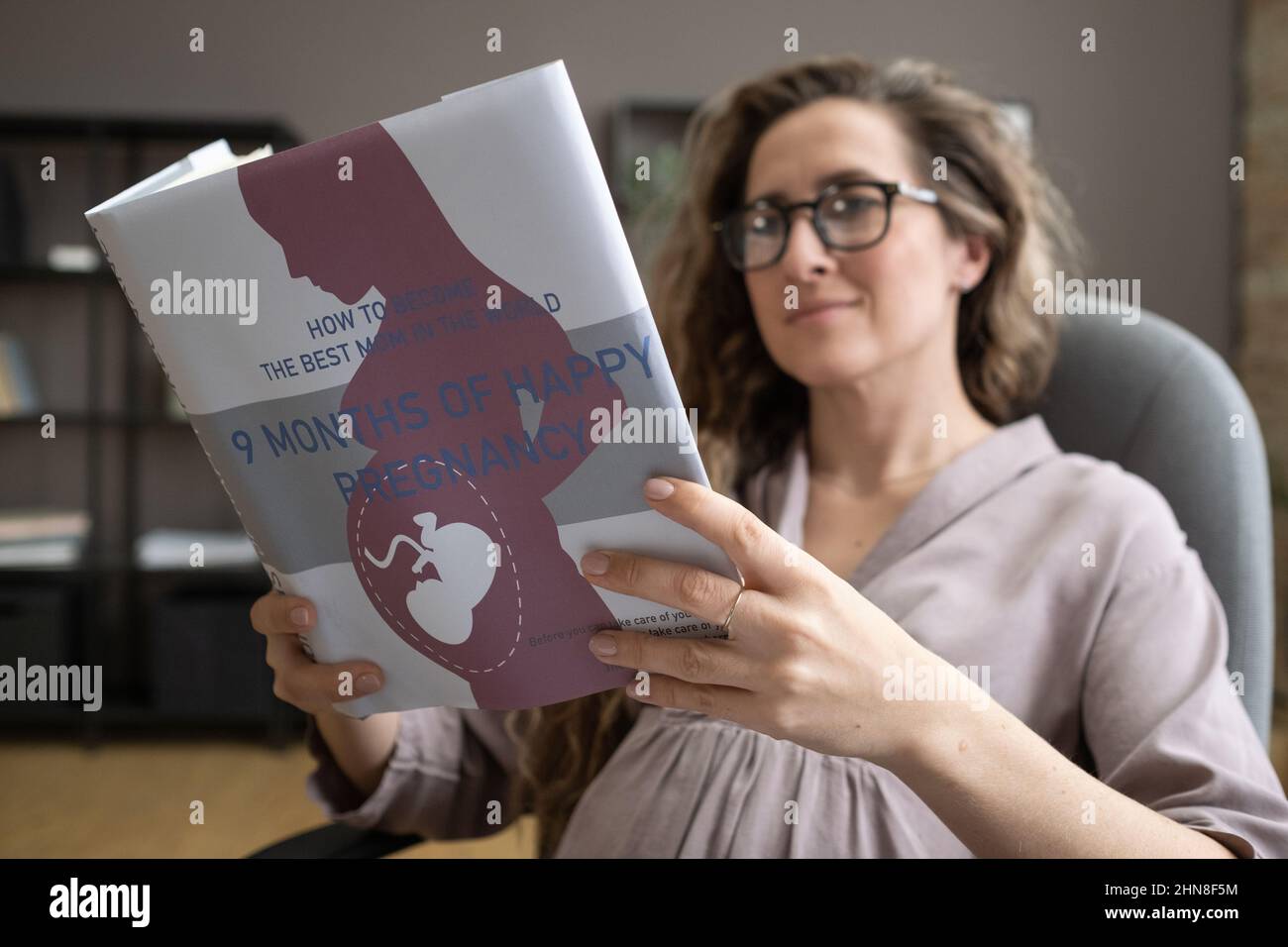 Jeune femme enceinte assise sur une chaise et lisant un livre sur la grossesse pendant qu'elle se repose après le travail au bureau Banque D'Images