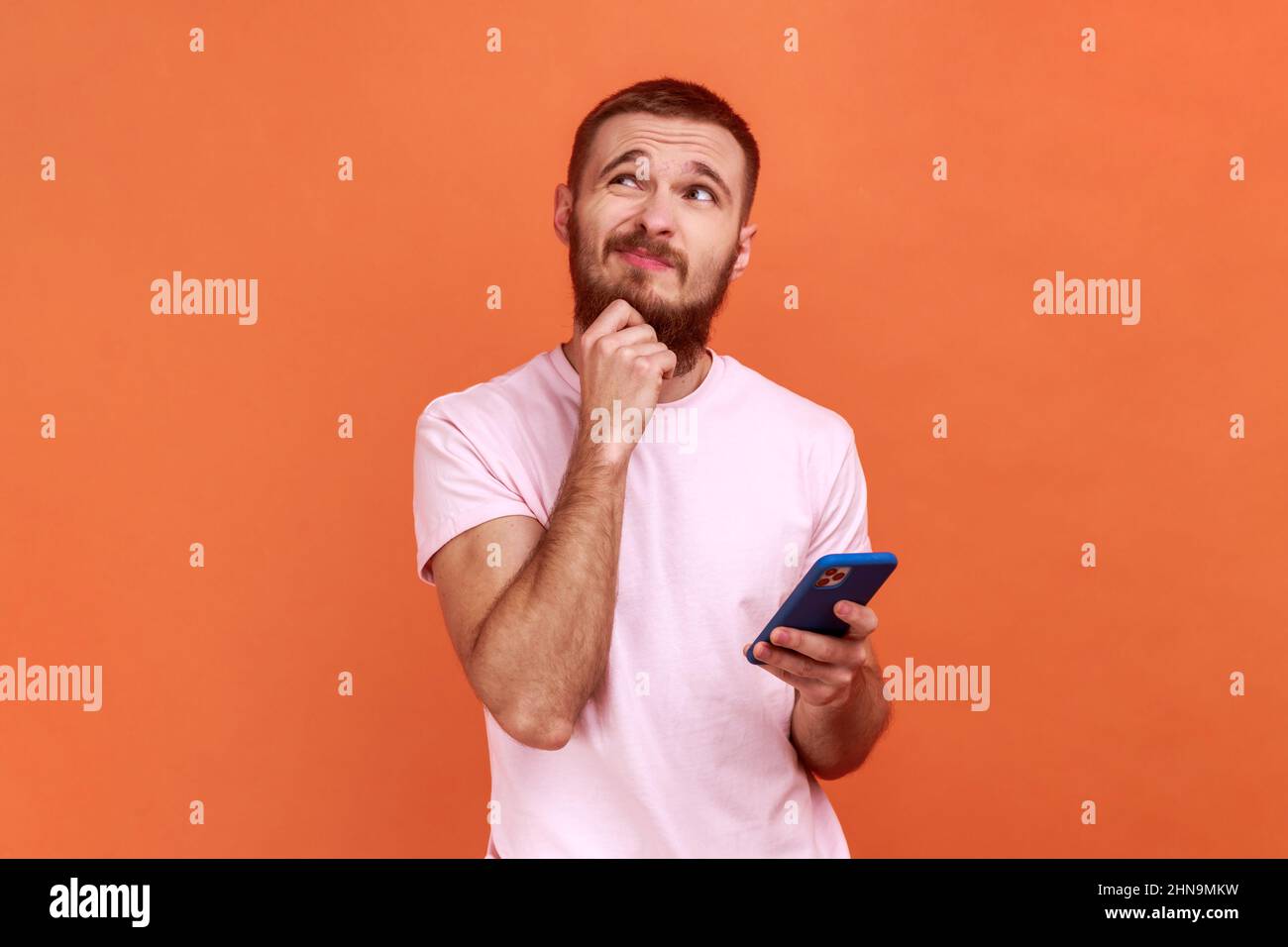 Portrait d'un homme tenant le menton, debout avec un smartphone à la main, contemplant la mise à jour du logiciel, choisissant les tarifs appropriés, portant un T-shirt rose. Studio d'intérieur isolé sur fond orange Banque D'Images
