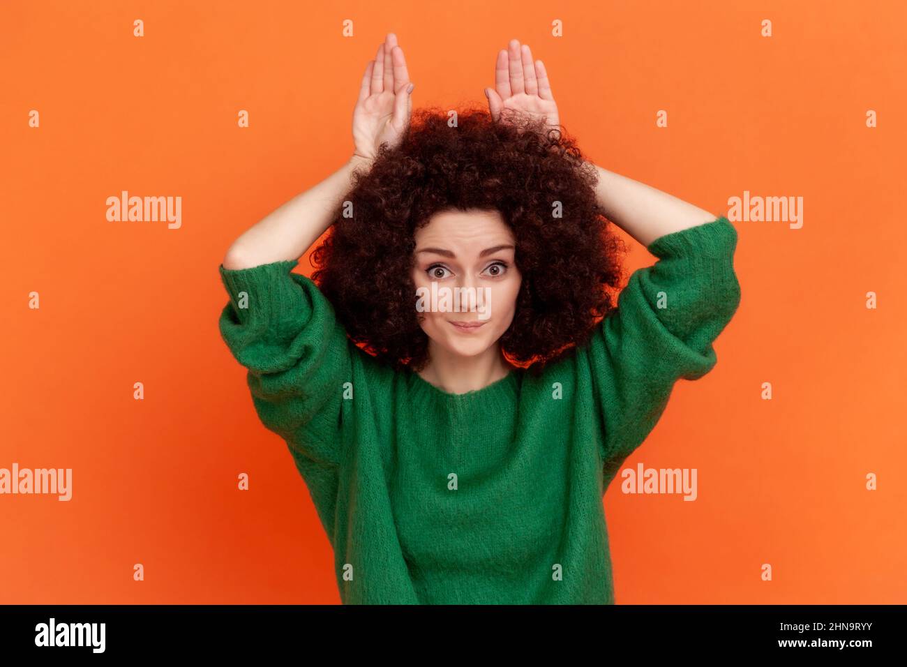 Femme avec une coiffure afro portant un pull vert décontracté montrant le geste des oreilles de lapin, tenant les mains sur la tête et souriant à l'appareil photo, s'amusant. Studio d'intérieur isolé sur fond orange. Banque D'Images