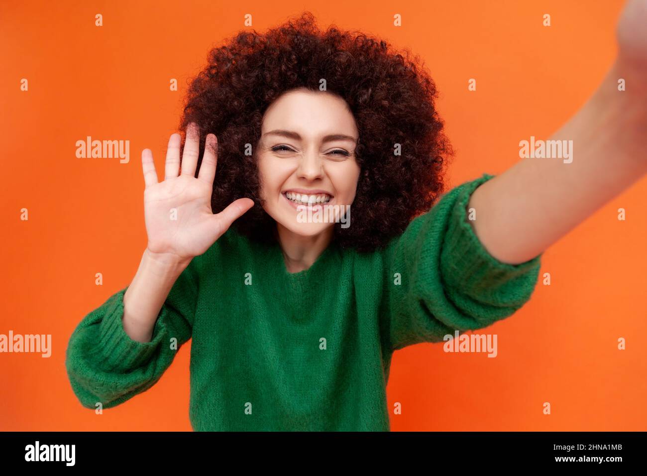 Une femme heureuse et satisfaite avec une coiffure afro portant un pull vert décontracté faisant selfie tourné par son smartphone, en agitant la main, en saluant ses adeptes. Studio d'intérieur isolé sur fond orange. Banque D'Images