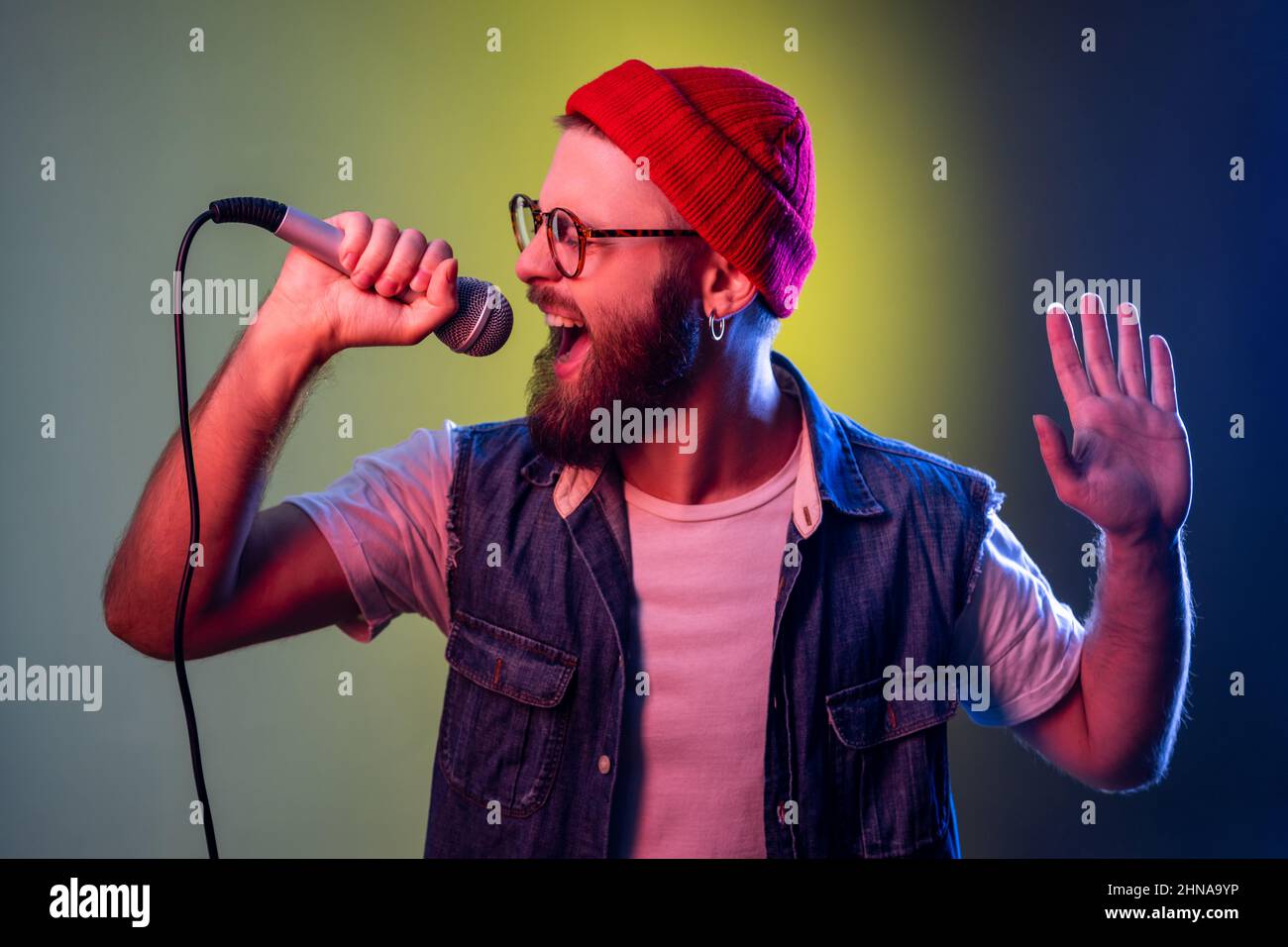 Un hipster passionné avec des chansons de barbe au karaoké tenant un microphone, chanteur de performance, de divertissement, a soulevé son bras. Studio d'intérieur isolé sur fond de néon coloré. Banque D'Images