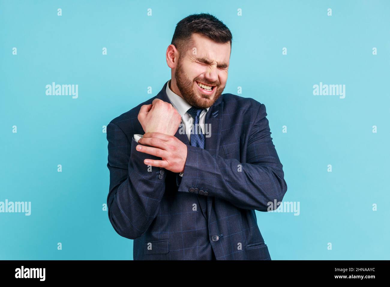Portrait d'un homme d'affaires malade portant un costume officiel debout avec de la grimace de douleur, massant mal au poignet, souffrant de blessure à la main ou d'entorse. Studio d'intérieur isolé sur fond bleu. Banque D'Images