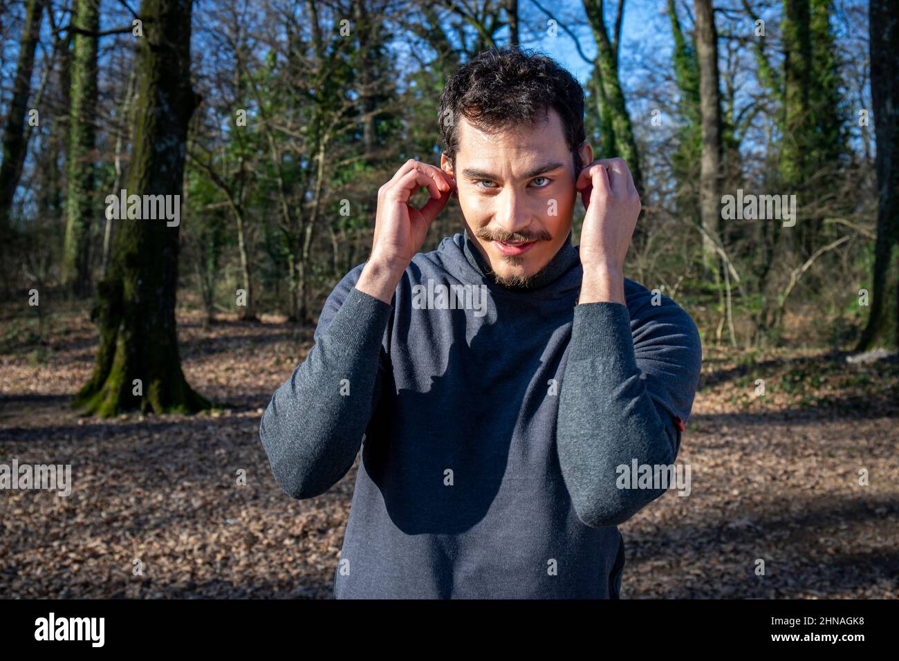Le coureur écoute de la musique tout en s'étirant dans les bois. Portrait d'un beau homme aux yeux bleus et d'une moustache qui s'entraîne dans le parc. Banque D'Images