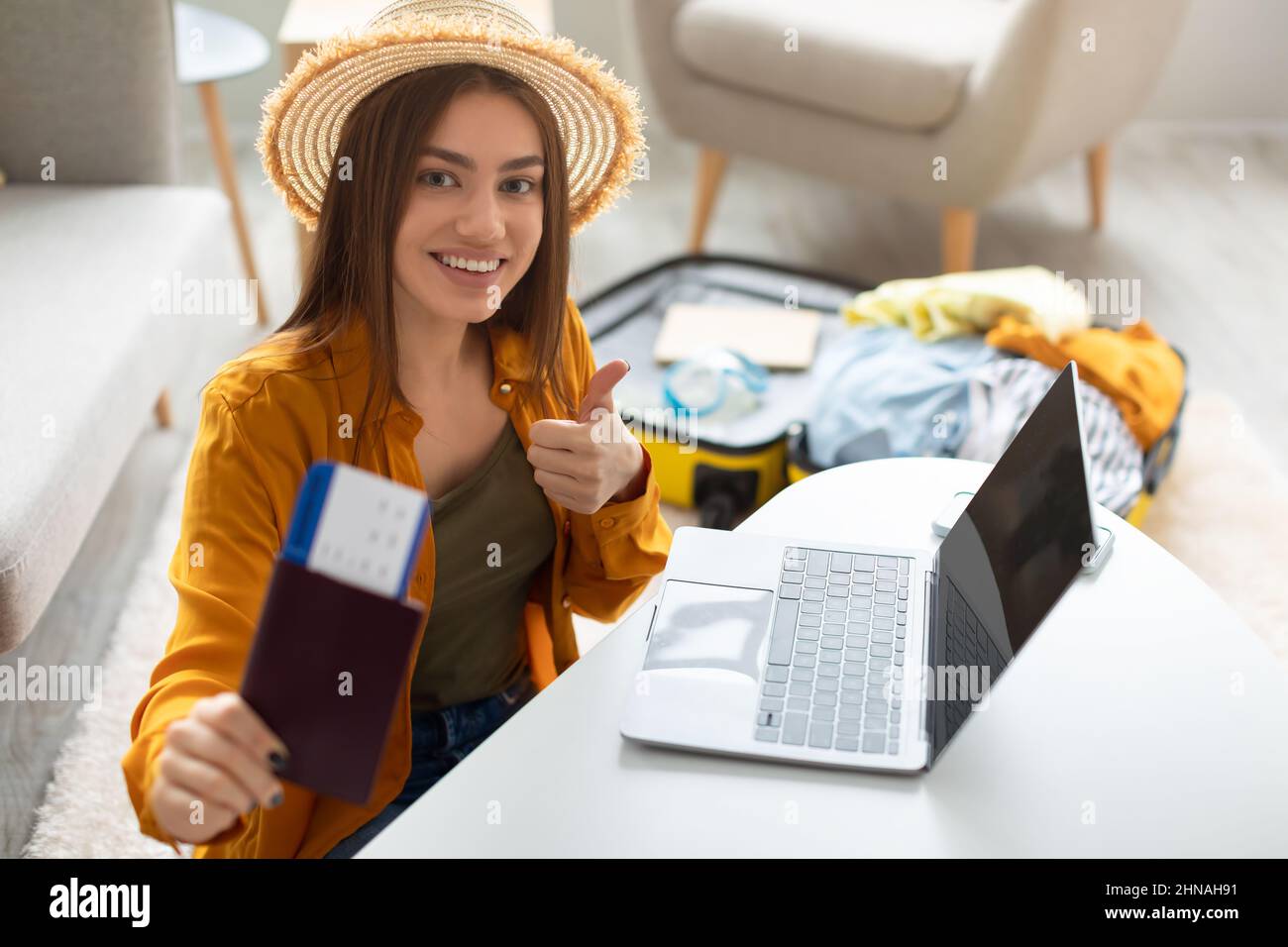 Une jeune femme heureuse avec un passeport et des billets à l'aide d'un ordinateur portable, montrant le pouce vers le haut, recommandant l'agence de voyage Internet à la maison Banque D'Images