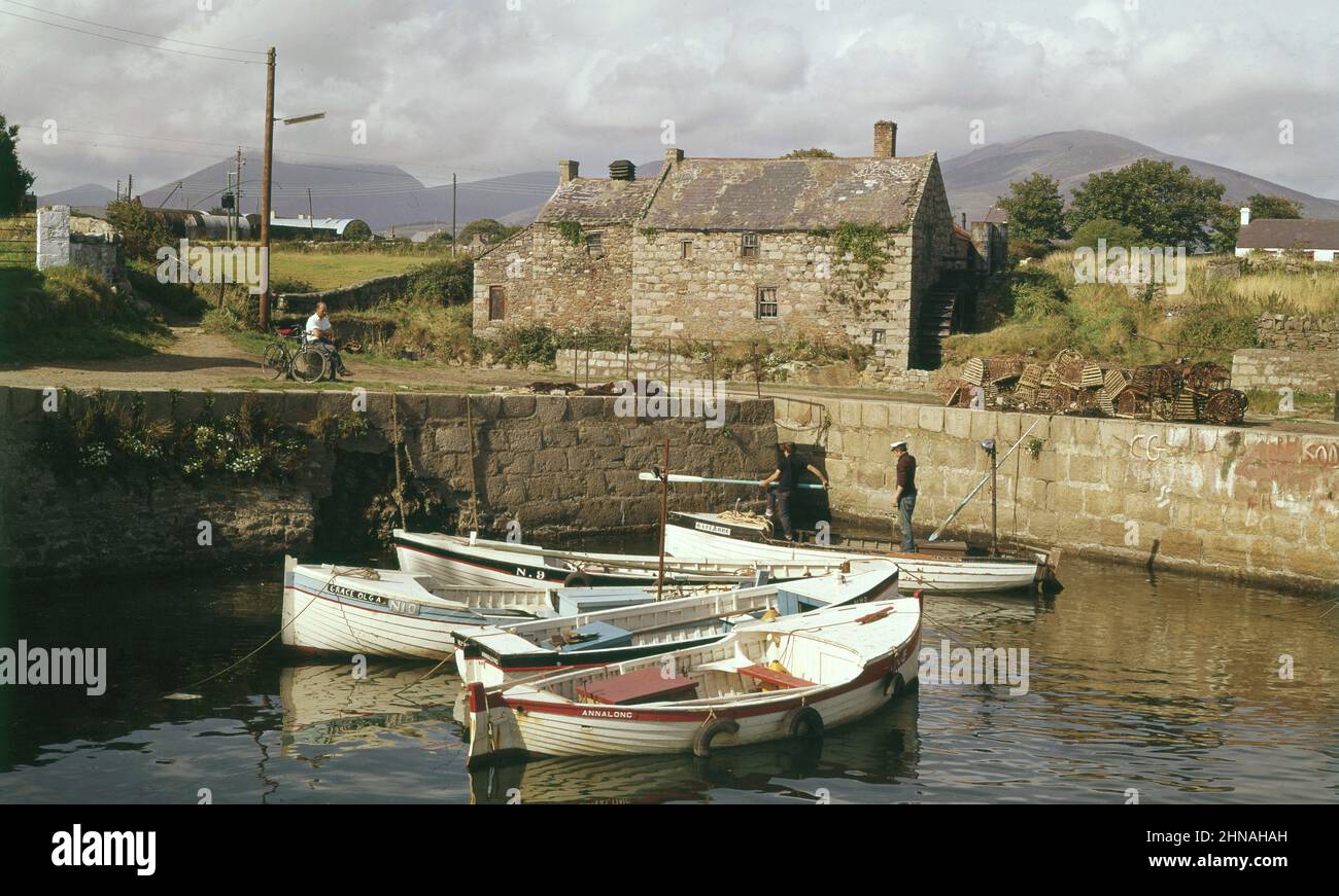 1960s, photo historique de cette époque de petits bateaux à rames amarrés dans le port d'Annalong, Co Down, Irlande du Nord, Royaume-Uni, avec deux lads debout en un et un homme local assis avec son vélo sur la terre sèche au-dessus. Village balnéaire pittoresque et paisible, Annalong se trouve au pied des montagnes de la Mourne. Banque D'Images