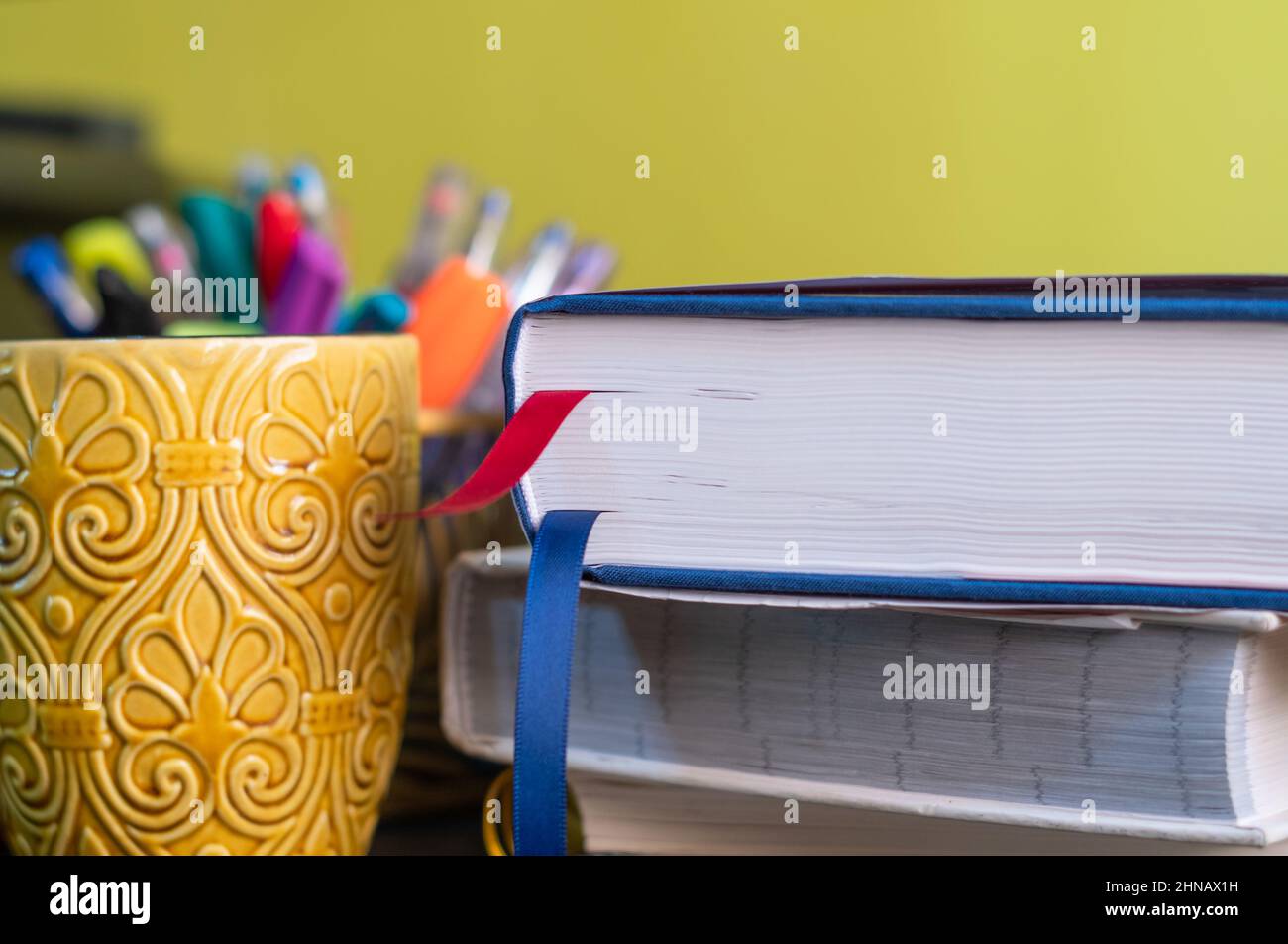 Bureau avec une pile de livres avec une tasse jaune et des stylos à l'arrière-plan. Banque D'Images