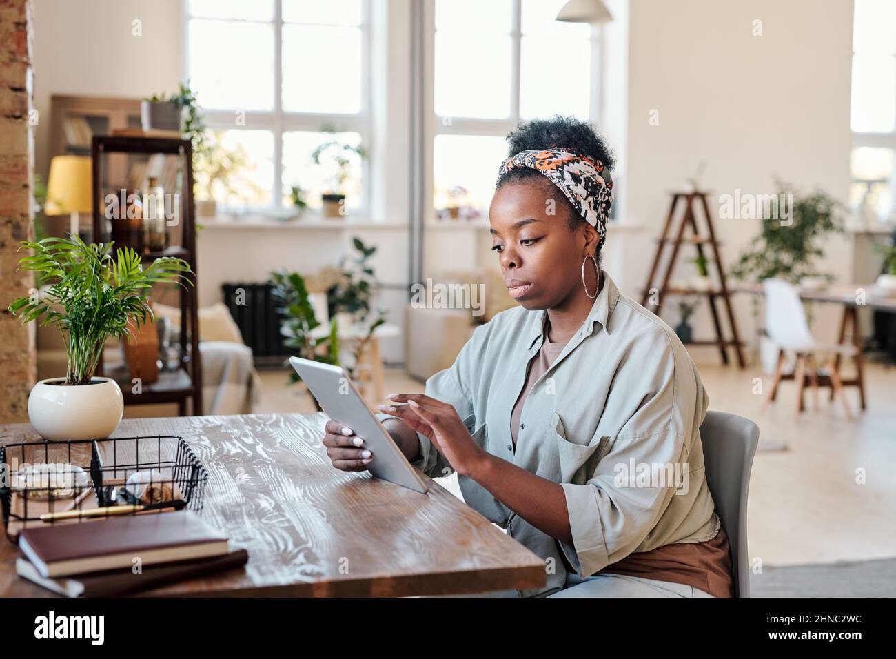 Jeune femme noire sérieuse en foulard assis sur un bureau en bois et travaillant sur un projet en ligne dans un bureau à espace ouvert Banque D'Images