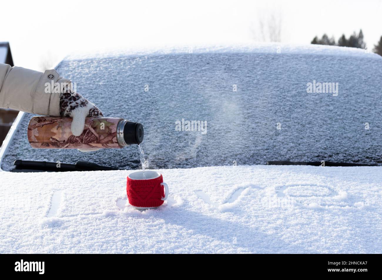 La main d'une femme dans une mitaine verse de l'eau bouillante dans une tasse d'un thermos sur une capuche enneigée d'une voiture lors d'une belle journée ensoleillée en hiver. L'inscription Banque D'Images